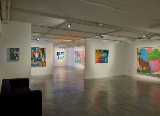 The image shows a contemporary gallery space with white walls displaying several colorful abstract paintings in various sizes. A dark purple bench is positioned in the foreground, and the room is well-lit with ceiling spotlights.