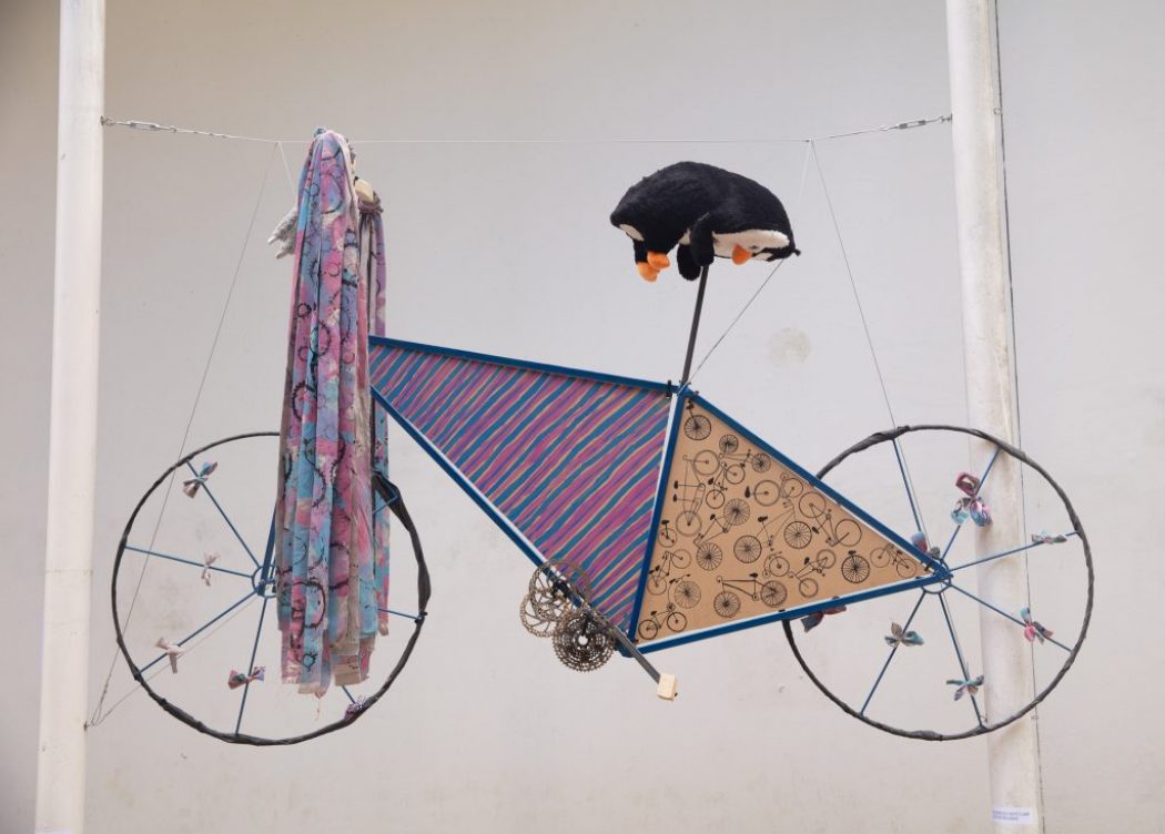 A sculptural piece resembling a bicycle is suspended, made from fabric, patterned panels, and a plush toy.