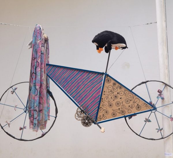 A sculptural piece resembling a bicycle is suspended, made from fabric, patterned panels, and a plush toy.