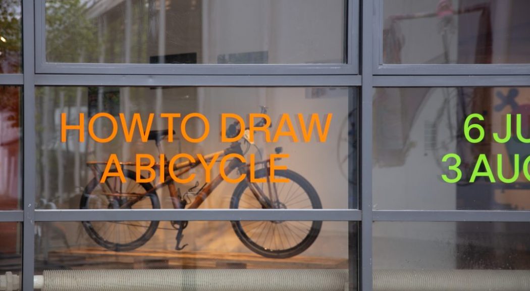A glass window displays orange and green text reading “HOW TO DRAW A BICYCLE” and exhibition dates, with a bicycle visible inside.