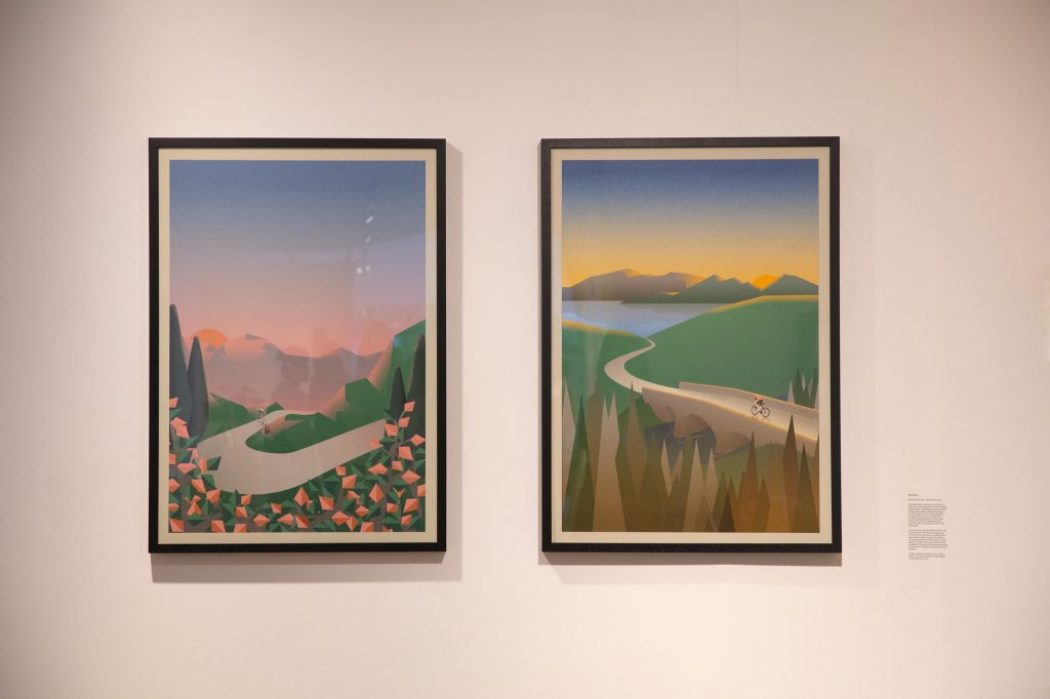 Two framed artworks hang on a gallery wall, each depicting stylized landscapes with winding roads and cyclists.