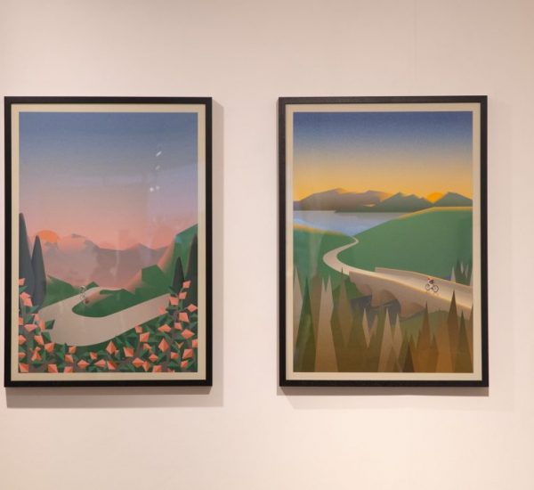 Two framed artworks hang on a gallery wall, each depicting stylized landscapes with winding roads and cyclists.