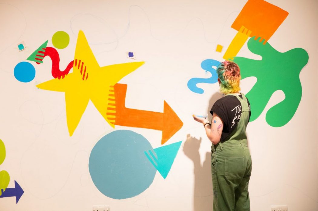A person stands in front of a wall painting colorful abstract shapes including stars, arrows, and circles.