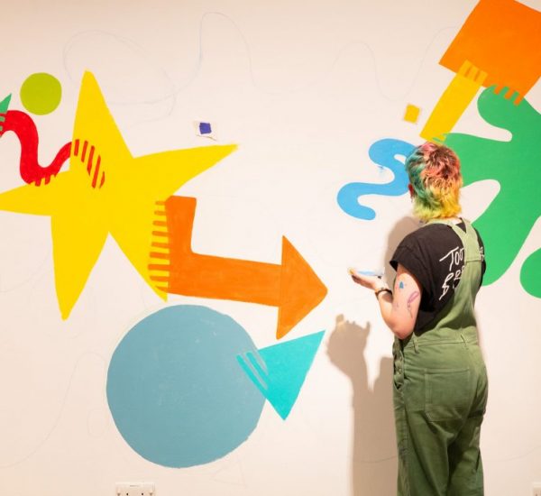 A person stands in front of a wall painting colorful abstract shapes including stars, arrows, and circles.