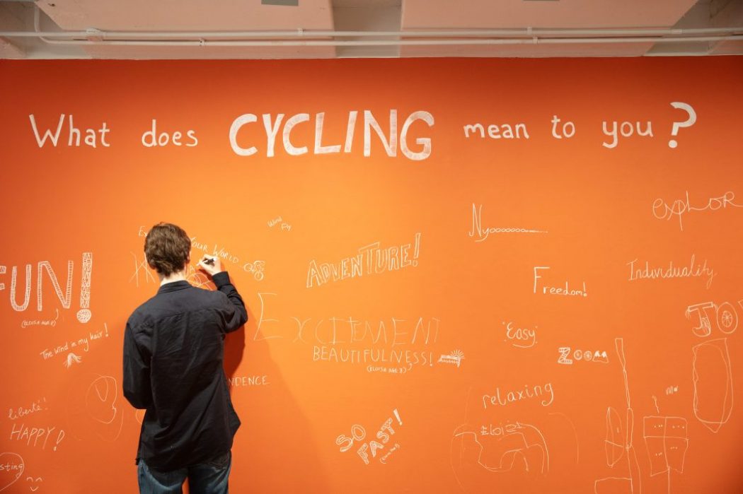 A person writes on a large orange wall with the question “What does CYCLING mean to you?” displayed in bold white letters. The wall is covered with handwritten responses such as “FUN!”, “Adventure!”, “Excitement,” and “Freedom.”
