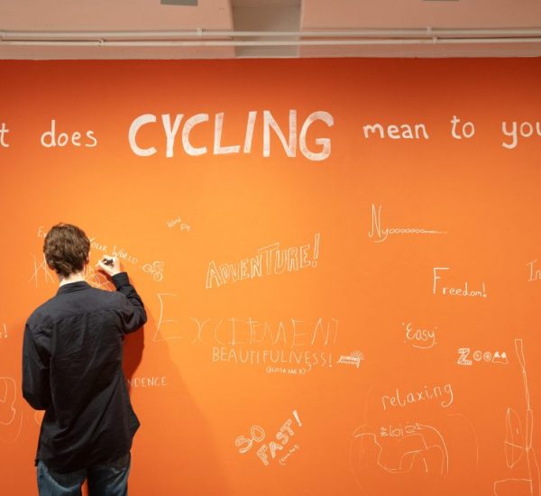 A person writes on a large orange wall with the question “What does CYCLING mean to you?” displayed in bold white letters. The wall is covered with handwritten responses such as “FUN!”, “Adventure!”, “Excitement,” and “Freedom.”
