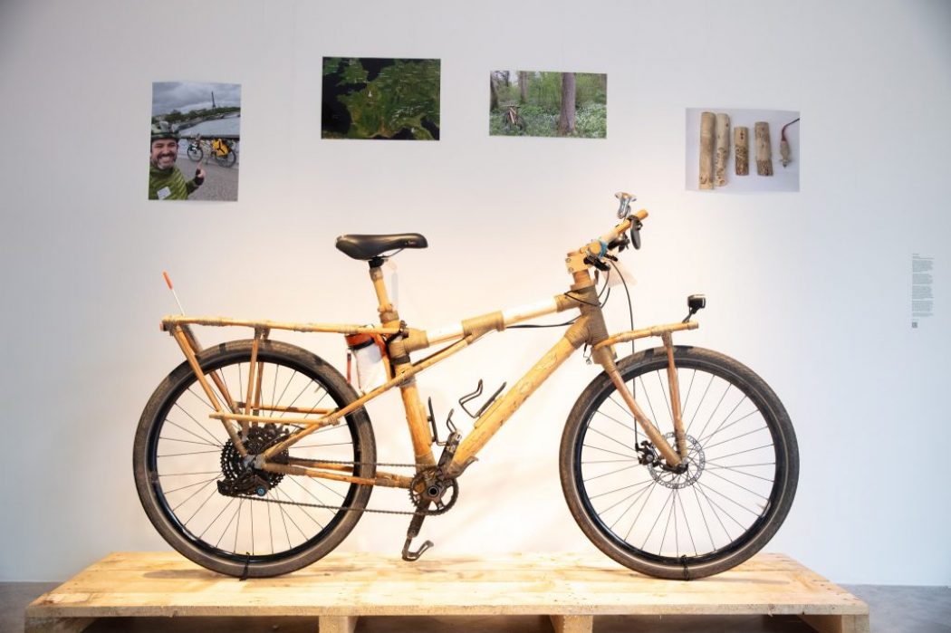 A bamboo-framed bicycle is displayed on a wooden pallet with photos and diagrams mounted on the wall behind it.