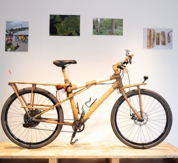 A bamboo-framed bicycle is displayed on a wooden pallet with photos and diagrams mounted on the wall behind it.