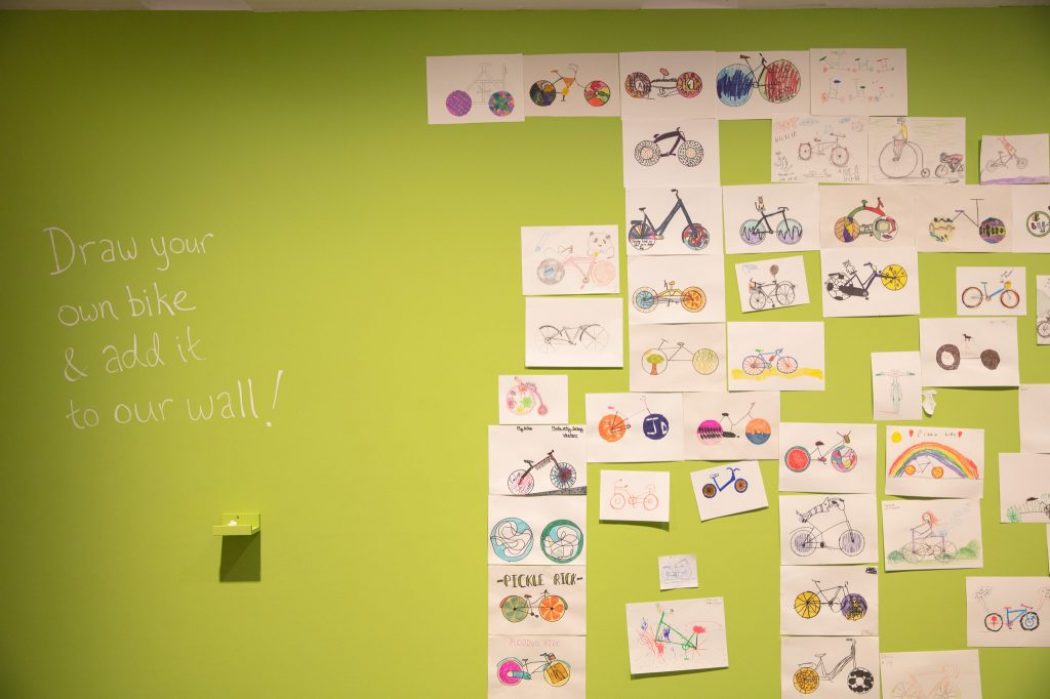 A green wall displays numerous hand-drawn bicycle illustrations arranged in a grid, with the text “Draw your own bike & add it to our wall!”.