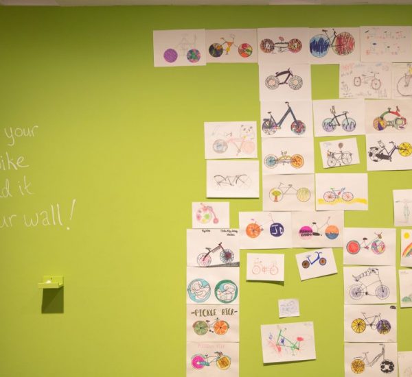 A green wall displays numerous hand-drawn bicycle illustrations arranged in a grid, with the text “Draw your own bike & add it to our wall!”.