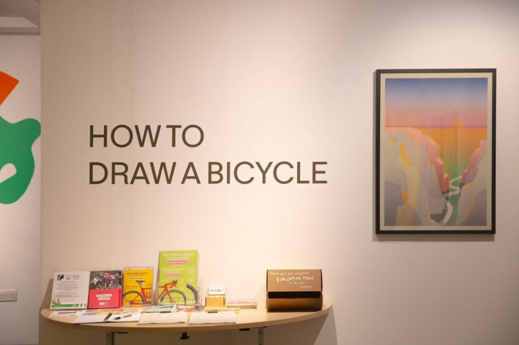 A gallery wall features the text “HOW TO DRAW A BICYCLE” above a table with leaflets and a framed artwork of a stylized landscape.