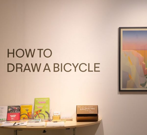 A gallery wall features the text “HOW TO DRAW A BICYCLE” above a table with leaflets and a framed artwork of a stylized landscape.