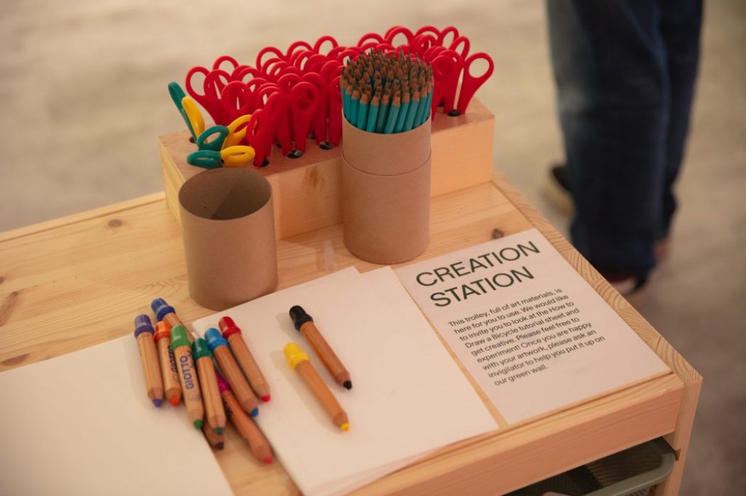A wooden table labeled “CREATION STATION” holds colored pencils, scissors, and blank paper for interactive activities.