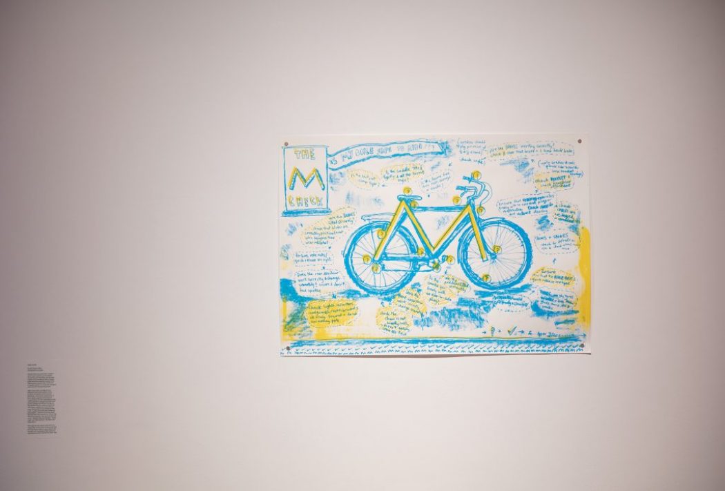 A hand-drawn artwork of a bicycle in blue and yellow is displayed on a white wall, surrounded by handwritten notes and sketches.