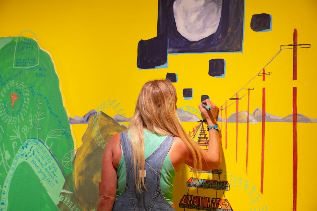 A person paints a bright yellow wall with colorful abstract shapes and lines, including telephone poles and a large dark square.
