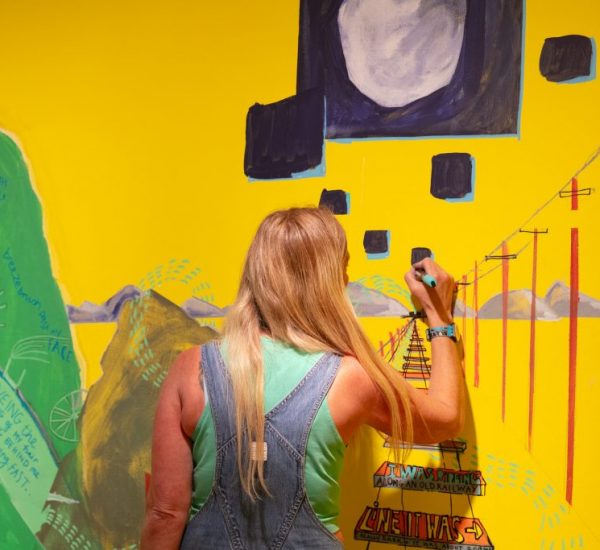 A person paints a bright yellow wall with colorful abstract shapes and lines, including telephone poles and a large dark square.