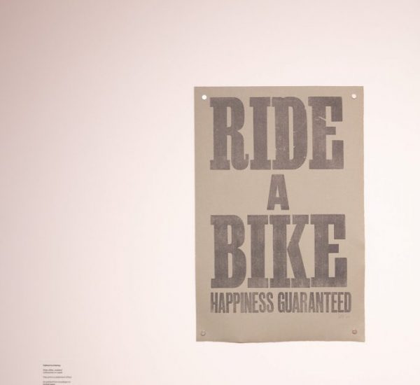 A poster with bold text reads “RIDE A BIKE HAPPINESS GUARANTEED” on a plain wall.