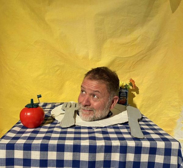 A head appears through a hole in a table covered with a blue-and-white checkered cloth, surrounded by props including a red apple-shaped object and cutlery shapes. A yellow backdrop with a sign reading “OPEN” hangs behind the setup.