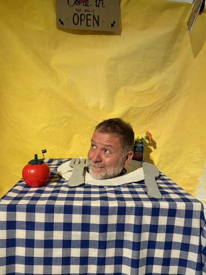 A head appears through a hole in a table covered with a blue-and-white checkered cloth, surrounded by props including a red apple-shaped object and cutlery shapes. A yellow backdrop with a sign reading âOPENâ hangs behind the setup.