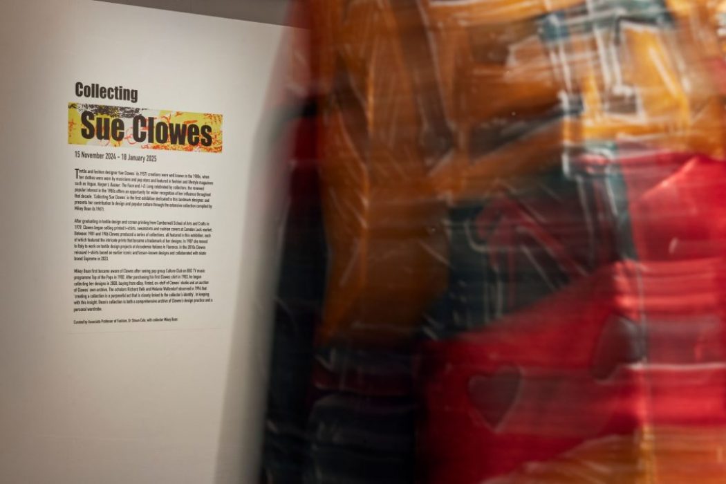 A gallery wall displays an introduction panel for the exhibition titled “Collecting Sue Clowes,” with colorful text and a detailed description. A blurred figure wearing vibrant patterned clothing is partially visible in the foreground.