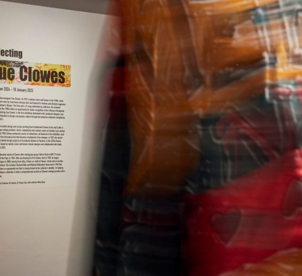 A gallery wall displays an introduction panel for the exhibition titled “Collecting Sue Clowes,” with colorful text and a detailed description. A blurred figure wearing vibrant patterned clothing is partially visible in the foreground.