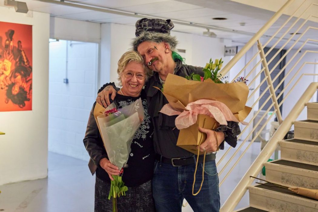Two people stand indoors holding large bouquets of flowers wrapped in paper, near a staircase and a wall with artwork. The setting appears to be a gallery or event space.