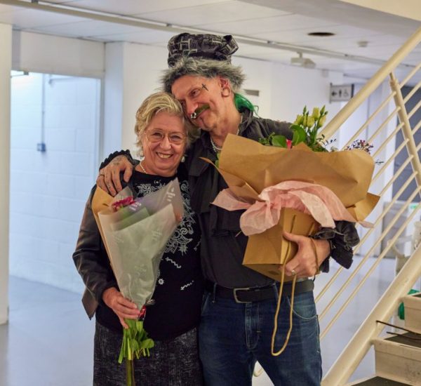 Two people stand indoors holding large bouquets of flowers wrapped in paper, near a staircase and a wall with artwork. The setting appears to be a gallery or event space.