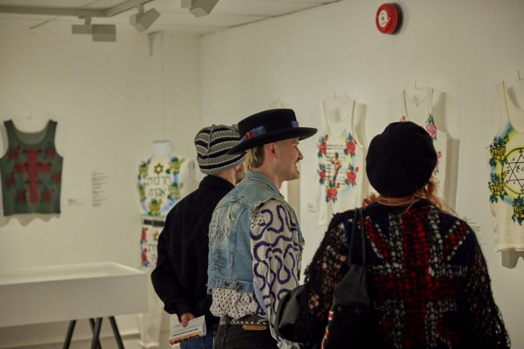 A gallery room features rows of mannequins dressed in patterned garments, creating a corridor-like view. Exhibition signage for “Collecting Sue Clowes” is visible in the background.