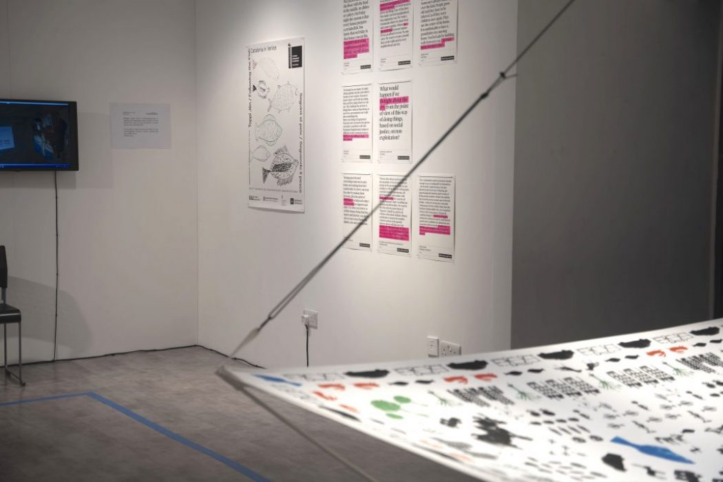 A gallery corner displays posters with black and pink text on white backgrounds, featuring design concepts and written content. In the foreground, part of a suspended fabric printed with various black and colored shapes is visible, and a monitor is mounted on the left wall.