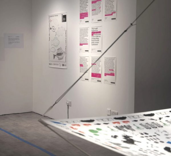 A gallery corner displays posters with black and pink text on white backgrounds, featuring design concepts and written content. In the foreground, part of a suspended fabric printed with various black and colored shapes is visible, and a monitor is mounted on the left wall.