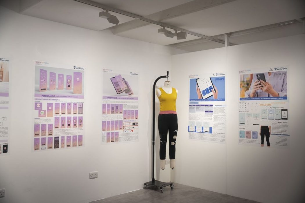 A gallery display features large posters on white walls showcasing app interface designs and informational graphics, primarily in purple and blue tones. In the center, a mannequin dressed in a yellow crop top and black leggings stands on a metal stand.