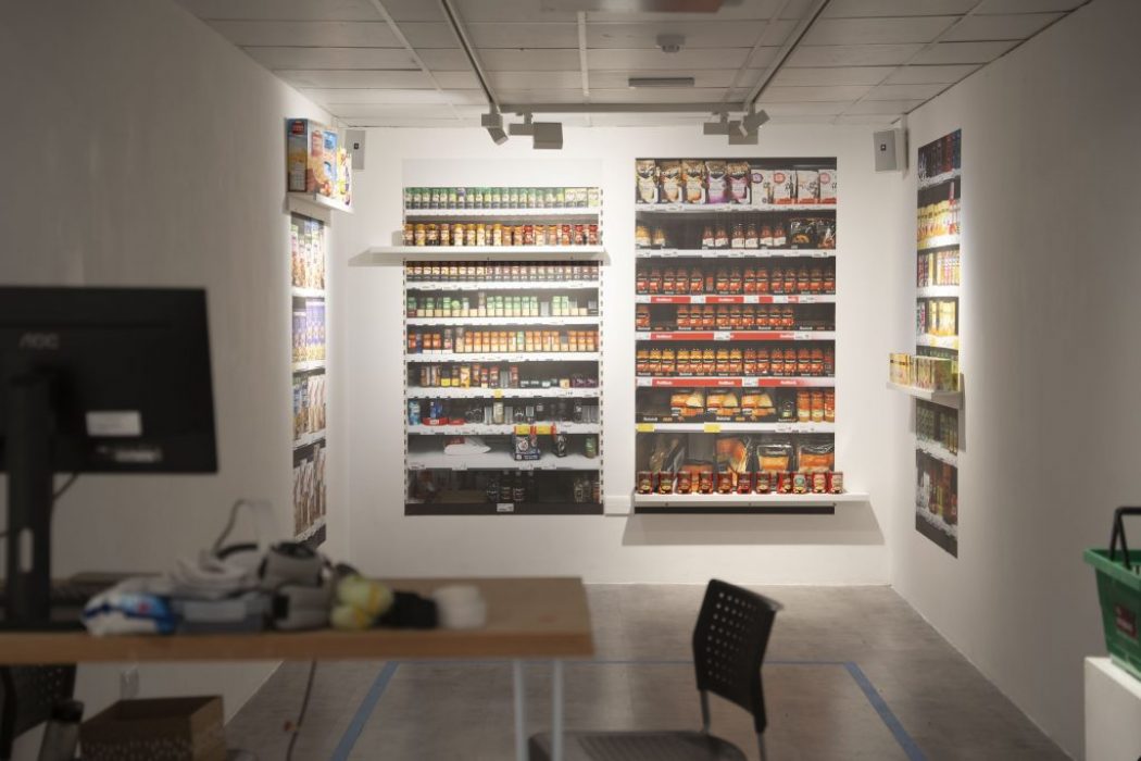 A room designed to resemble a small grocery store features shelves lined with colorful packaged goods, including cereals and canned items, on three walls. In the foreground, there is a desk with a monitor and VR equipment, and a black chair facing the display.