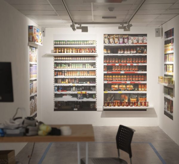 A room designed to resemble a small grocery store features shelves lined with colorful packaged goods, including cereals and canned items, on three walls. In the foreground, there is a desk with a monitor and VR equipment, and a black chair facing the display.