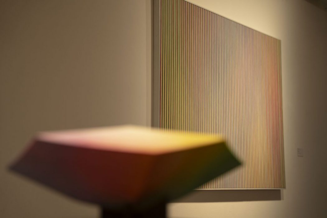A blurred gradient-colored sculpture in the foreground contrasts with a background painting of vertical multicolored lines.