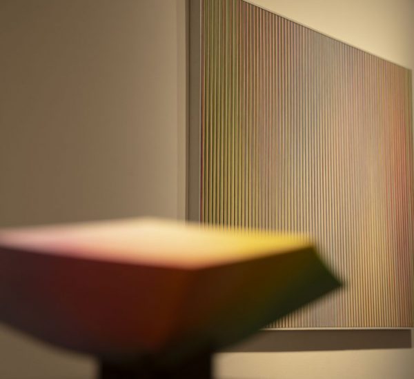 A blurred gradient-colored sculpture in the foreground contrasts with a background painting of vertical multicolored lines.