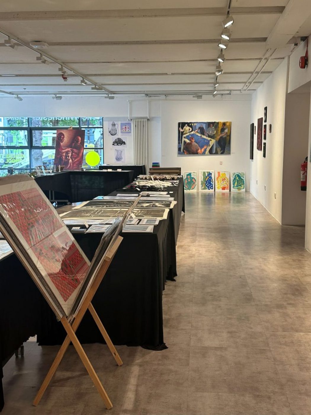 An art gallery with tables covered in black cloth displaying prints and artworks, with a wall in the background featuring a large painting and four smaller portraits.