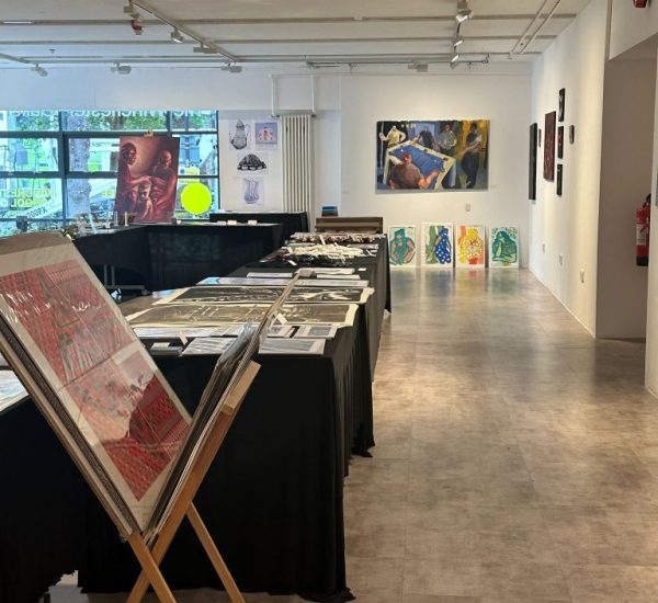 An art gallery with tables covered in black cloth displaying prints and artworks, with a wall in the background featuring a large painting and four smaller portraits.