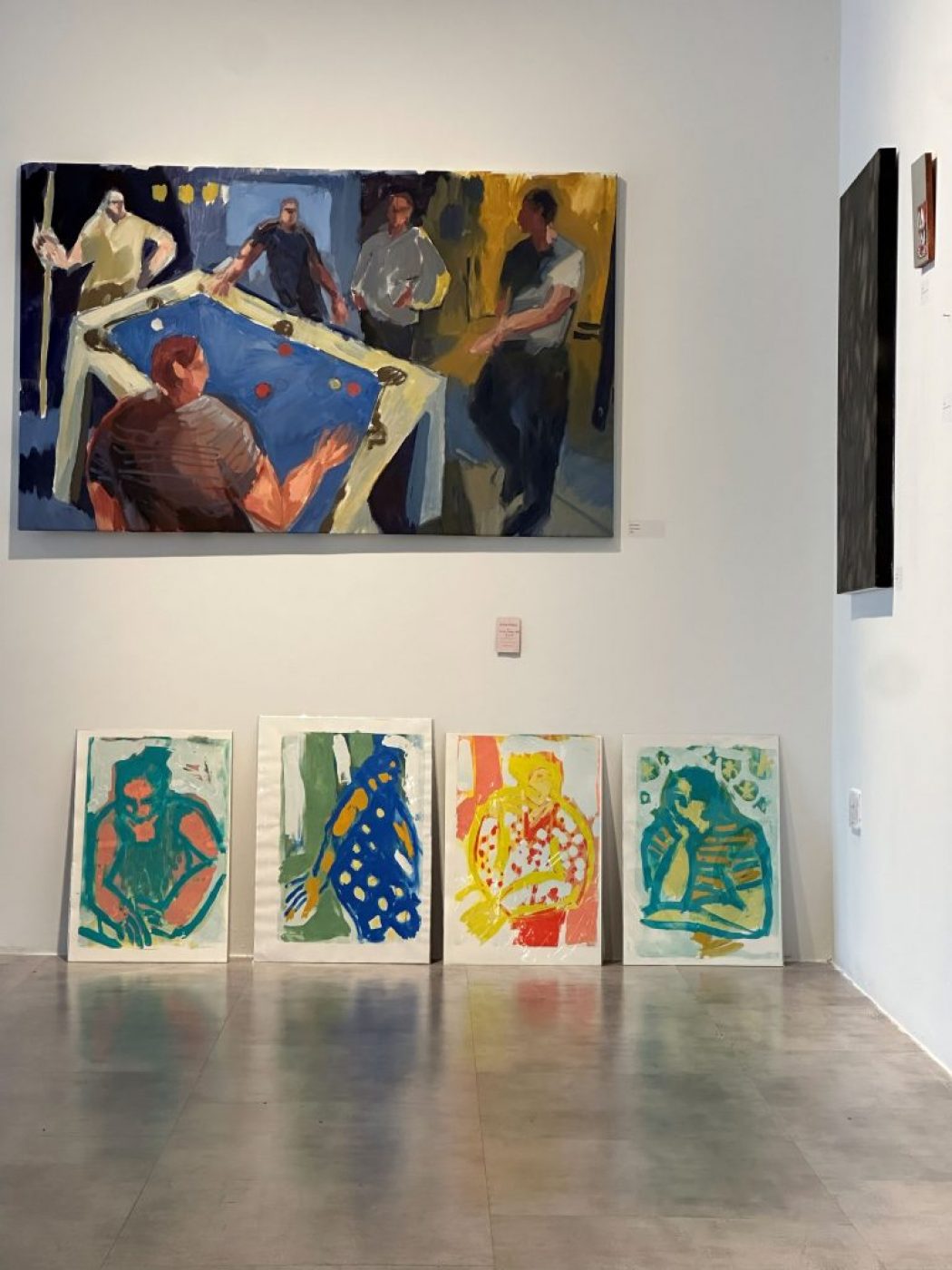 A large painting of people around a pool table hangs on a white wall, with four colorful abstract portraits displayed on the floor beneath it.
