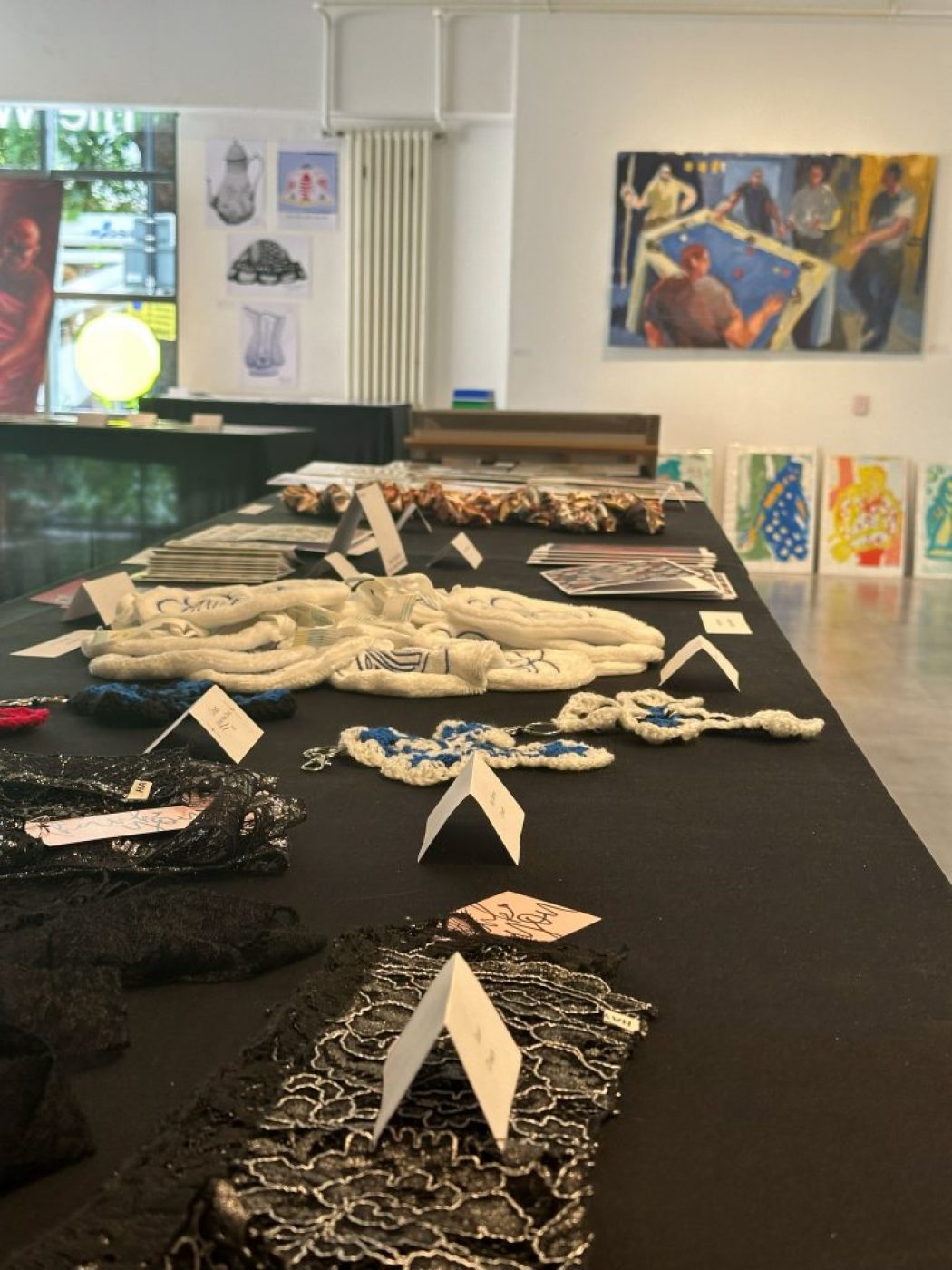 A close-up view of a display table with handmade textile pieces and jewelry, with a large painting and colorful portraits visible in the background.