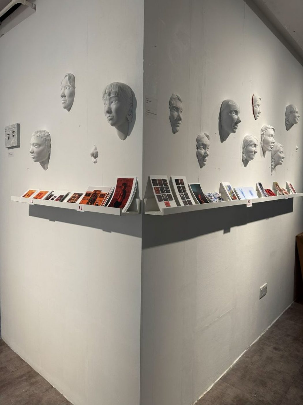 White sculpted faces are mounted on two adjoining walls above shelves displaying small colorful prints and booklets.