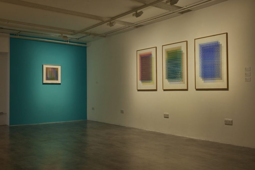 A gallery room features three framed abstract artworks with intricate patterns on one wall and a single striped artwork on a teal-colored wall.