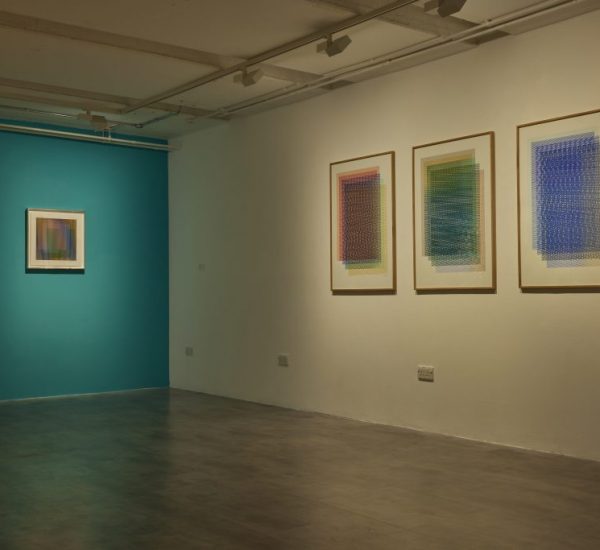 A gallery room features three framed abstract artworks with intricate patterns on one wall and a single striped artwork on a teal-colored wall.