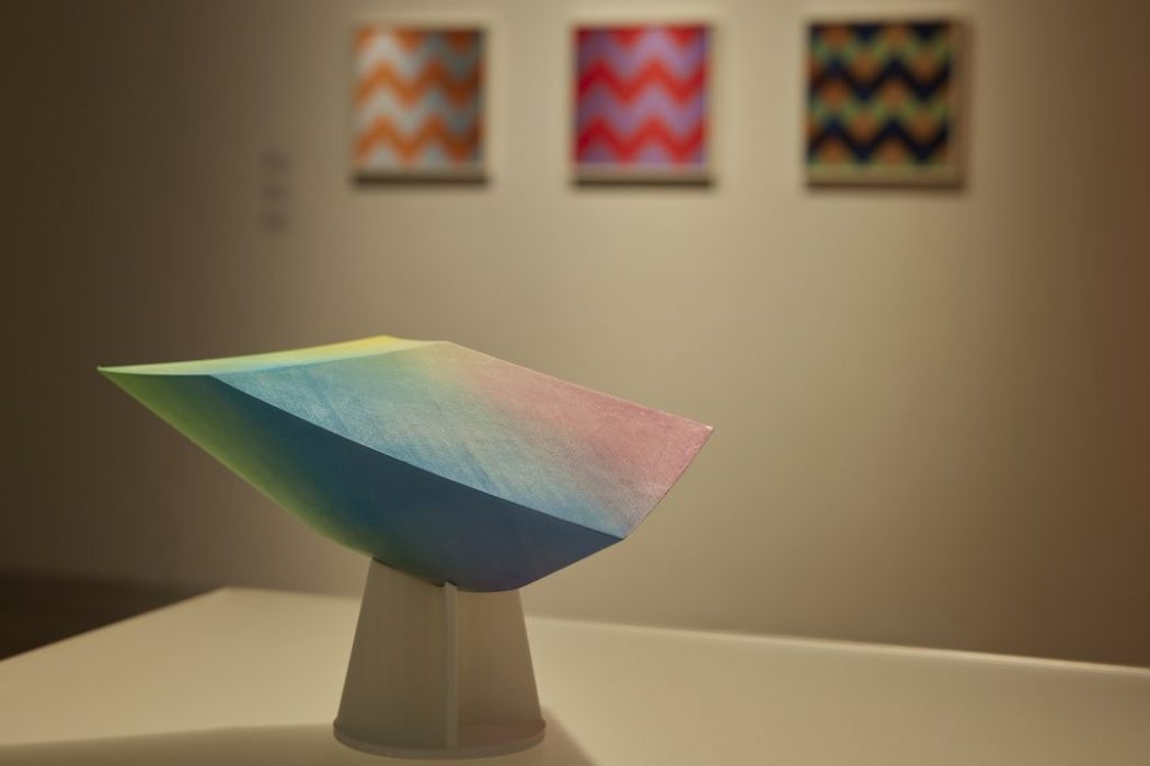 A colorful geometric sculpture stands on a pedestal in front of three vibrant zigzag-patterned paintings in an art gallery.