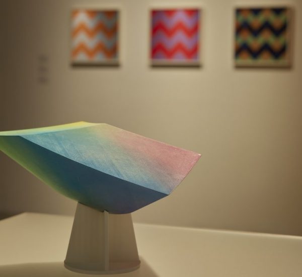 A colorful geometric sculpture stands on a pedestal in front of three vibrant zigzag-patterned paintings in an art gallery.