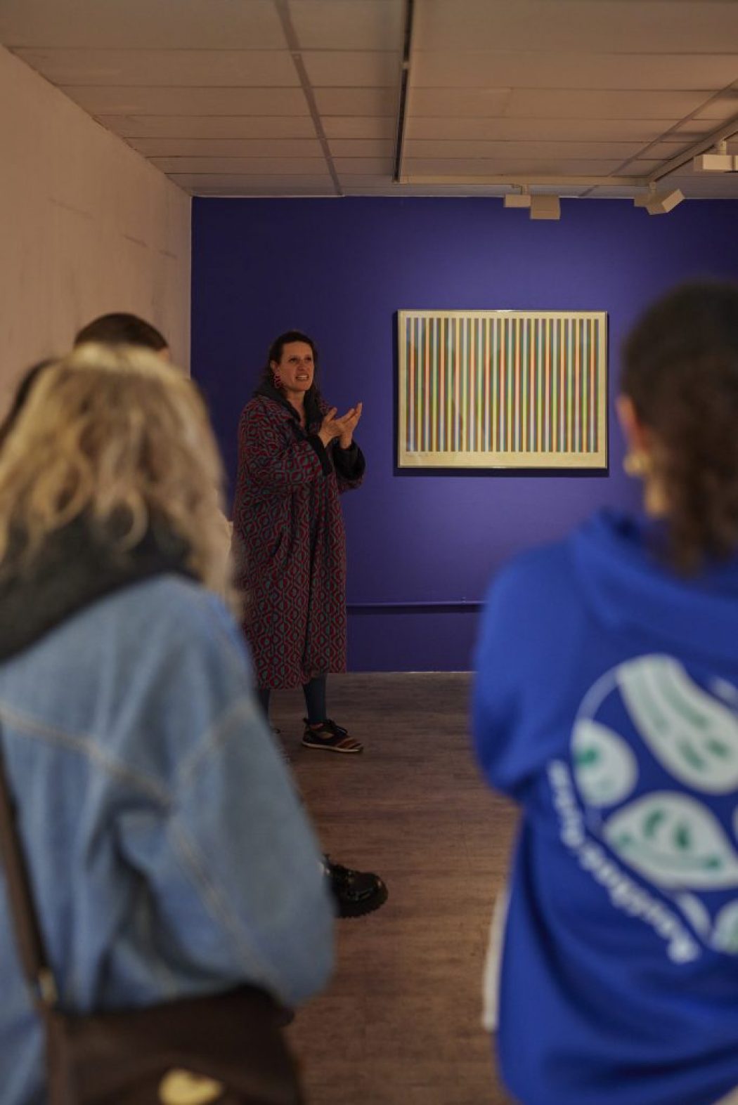A group of people listens to a speaker in front of a striped abstract painting mounted on a purple accent wall in an art gallery.
