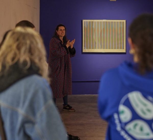 A group of people listens to a speaker in front of a striped abstract painting mounted on a purple accent wall in an art gallery.
