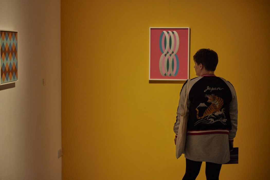 A person observes an artwork of interlocking loops on a yellow wall, with another geometric painting visible on an adjacent white wall.