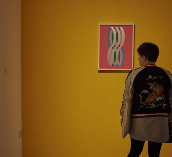 A person observes an artwork of interlocking loops on a yellow wall, with another geometric painting visible on an adjacent white wall.