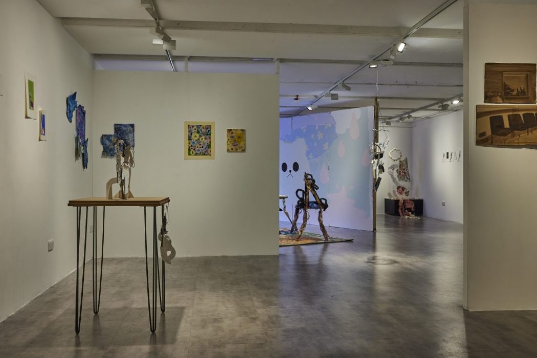 A contemporary art gallery with white walls displays various colorful paintings and mixed-media artworks, including a wooden table sculpture in the foreground. In the background, a large wall mural with abstract shapes and a face-like design is visible, along with other sculptures and hanging pieces.