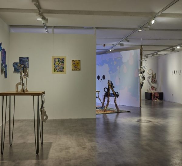 A contemporary art gallery with white walls displays various colorful paintings and mixed-media artworks, including a wooden table sculpture in the foreground. In the background, a large wall mural with abstract shapes and a face-like design is visible, along with other sculptures and hanging pieces.